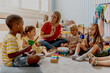 © lordn - Preschool teacher leading a fun interactive activity with children sitting in a circle, raising hands and smiling in a bright classroom
