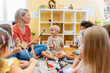 © lordn - Preschool teacher leading a fun interactive activity with children sitting in a circle, raising hands and smiling in a bright classroom