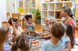 © lordn - Preschool teacher leading a fun interactive activity with children sitting in a circle, raising hands and smiling in a bright classroom