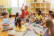 © lordn - Preschool teacher leading a fun interactive activity with children sitting in a circle, raising hands and smiling in a bright classroom