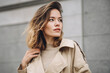 © EricMiguel - Stylish young woman in a beige trench coat posing confidently outdoors with soft natural lighting