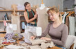 © JackF - Focused on stitching, skilled young female seamstress sewing linen fabric at sewing machine in small workshop, while colleague working on fitting dress to mannequin in background
