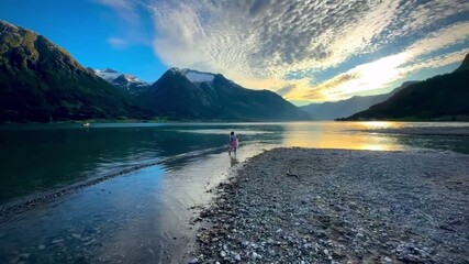 Wall Mural - girl in a pink dress runs joyfully along a rocky shore by a calm lake at sunset