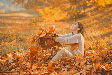 Wall Mural - Girl in autumn orange leaves. Yellow fallen maple leaf holds a female hand in the park. Portrait of beautiful young woman outdoors in autumn. Large maple leaf yellow orange and brown in a female hand.