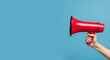 © Dionisia - Hand holding a red and black megaphone on a blue background. Ideal for messages, announcements, protests, or communication.