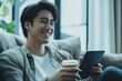 © Ifti Digital - Smiling Asian man relaxing on couch while enjoying coffee and using tablet at home during daytime, Smiling Asian man watching something on tablet and drinking coffee at home