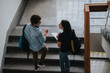 © qunica.com - Two university students engage in a lively conversation while standing on a staircase. Both carry backpacks and one holds a notebook, indicating a study setting.