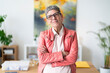 © Studio Marmellata - Middle aged woman in glasses and pink leather jacket crossing arms and looking at camera while standing in modern office
