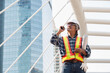 © AU USAnakul - Southeast Asian construction engineer in safety helmet and reflective vest holding blueprint while using walkie-talkie at modern urban site, supervising smart city infrastructure development.