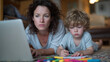© Maksym - A tired mother leans forward toward her laptop, face showing strain as she juggles deadlines. Next to her, a child draws with vibrant markers on the floor, immersed in creativity.