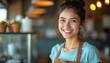 © Maryna - Portrait of smiling young woman wearing apron in cafe or shop. She has dark hair tied up, appears friendly, confident. Background is softly blurred, suggesting cozy, inviting atmosphere for customers.
