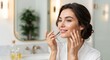 © Elitemedia - A beautiful young woman with a radiant smile applies moisturizing cream to her face while looking in the mirror. Skincare routine and self-care concept in a modern bathroom.