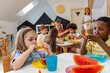 © lordn - Diverse preschool children enjoying fresh watermelon at a table in a bright classroom, developing healthy eating habits and social skills during snack time
