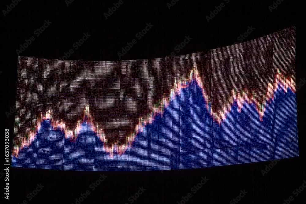 A large digital screen shows a stock market trend with peaks and valleys prominently displayed. The colors blue and pink contrast against a dark background, depicting fluctuations over time.
