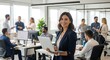 © Ray - Confident woman in business attire holds a laptop in a modern office. Coworkers are visible in the background. She smiles, projecting leadership & success.