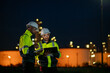 © APchanel - Engineers wearing safety gear, including hard hats examining survey are using tablet collaborating and discussing on screen work plant site use with night lights of oil refinery industry background