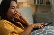 © Johnstocker - Side view, Middle-aged Asian woman relaxes on the sofa, looking drowsy while watching a movie in the living room at home.