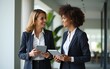 © fu - Two businesswoman working together, while walking in office and holding digital gadget pc. Small creative diversity team of African American and blond females executives meeting discuss work project.