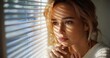 © Gzvzattio - Thoughtful woman with blond hair looking through window blinds with soft natural light casting striped shadows on her face expressing contemplation and calmness