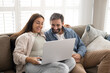© fizkes - Happy young couple sitting on comfortable couch in living room, browsing internet, buying goods and electronic services together, watching movie or TV show using modern laptop. Marketplace, e-commerce