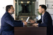 © fizkes - Two diverse male younger and older business coworkers arguing at work table, sitting opposite, talking, having conflict of generations. Senior boss dissatisfied with young professional paper report