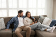 © fizkes - Middle-aged husband hugs young wife sitting together on sofa, relaxing, talking, spend pastime on internet using smartphone, checking e-bank, making purchases via marketplaces application. Rest, tech