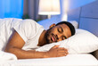 © theshooter - Un hombre joven dormido descansando durante la noche en una cama con sabanas y almohada blanca con la luz del dormitorio encendida