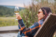 © EdNurg - Young woman sitting on wooden chair, holding glass of white wine, enjoying landscape in vineyard