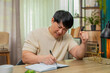 © Andrii Iemelianenko - Asian man sitting at home table writes budget notes, crossing numbers again, rewriting lines. Chinese guy frowns, tapping pen, showing stress while planning financial decision and seeking solution.