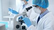 © rogerphoto - Masked scientist examining specimen under microscope while laboratory assistant handles glassware in a professional medical investigation. Health care and science concept