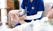 © NAMPIX - Nurse in blue scrubs checking pulse of senior male patient during home visit, close-up of hands.