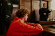 © AnnaStills - Caucasian teenage boy with curly hair sitting in courtroom wearing orange jumpsuit, resting arms on wooden barrier, security officer and male judge standing in background during trial