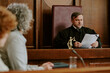 © AnnaStills - Caucasian middle aged man judge reading legal documents in courtroom while two women listening in foreground, legal proceedings taking place in judicial setting with focus on authority