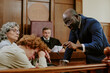 © AnnaStills - Black middle aged man in suit addressing Caucasian teenager with head down while middle aged Caucasian woman and judge, observing in courtroom setting during legal proceedings