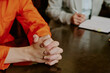 © AnnaStills - Caucasian young adult man sitting at table with hands clasped in front, wearing orange jumpsuit, legal professional writing in notebook during meeting or consultation