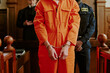 © AnnaStills - Caucasian young adult man standing in courtroom wearing handcuffs and orange jumpsuit with hands in front, police officer and judge visible in background during legal proceedings