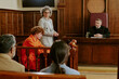 © AnnaStills - Caucasian teenage boy sitting in defendant box wearing orange jumpsuit, middle aged Caucasian woman standing and speaking, middle aged Caucasian male judge listening in courtroom