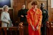 © AnnaStills - Caucasian teenage boy with curly hair standing in handcuffs wearing orange jumpsuit, guarded by Black male police officer in courtroom with middle aged female lawyer and male judge observing