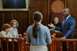 © AnnaStills - Black middle aged man in suit addressing Caucasian young adult woman standing in witness box, while Caucasian middle aged woman and Caucasian young adult man observing in courtroom
