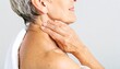© BROPRADANA - An older woman with short gray hair, draped in a white towel, gently touches her neck with her right hand.