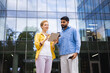 © sofiko14 - Two business professionals are reviewing a tablet outside a modern office building on a sunny day.