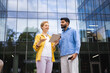 © sofiko14 - Two business colleagues discuss on a tablet outdoors in front of a modern office building.