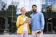 © sofiko14 - Two diverse business colleagues stand in front of a modern office building, smiling and looking at the camera, one holding a tablet.