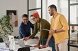 © TrueFrame Collective - Medium long shot of three cheerful young and mature Indian men working on business project in modern office