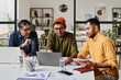 © TrueFrame Collective - Group of three Indian men sitting at table in modern office working on business project, looking at laptop screen and discussing plan
