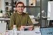 © TrueFrame Collective - Medium portrait of young Indian man wearing casual clothes holding digital tablet sitting at table in office smiling at camera