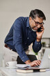 © TrueFrame Collective - Vertical medium long shot of cheerful South Asian businessman talking on phone and checking data on laptop