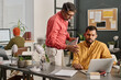© TrueFrame Collective - Mature Indian man holding digital tablet standing at desk in modern office looking at laptop screen and helping his younger co-worker