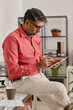 © TrueFrame Collective - Vertical medium long shot of mature indian man sitting on desk in modern office making notes on digital tablet with use of stylus