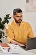 © TrueFrame Collective - Vertical shot of serious young Indian man in yellow shirt sitting at desk in modern office typing on laptop
